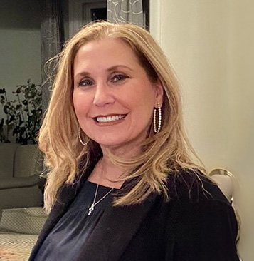The image shows a woman posing for a portrait, wearing a dark top with a light-colored blazer over it, accompanied by a smile and looking directly at the camera.
